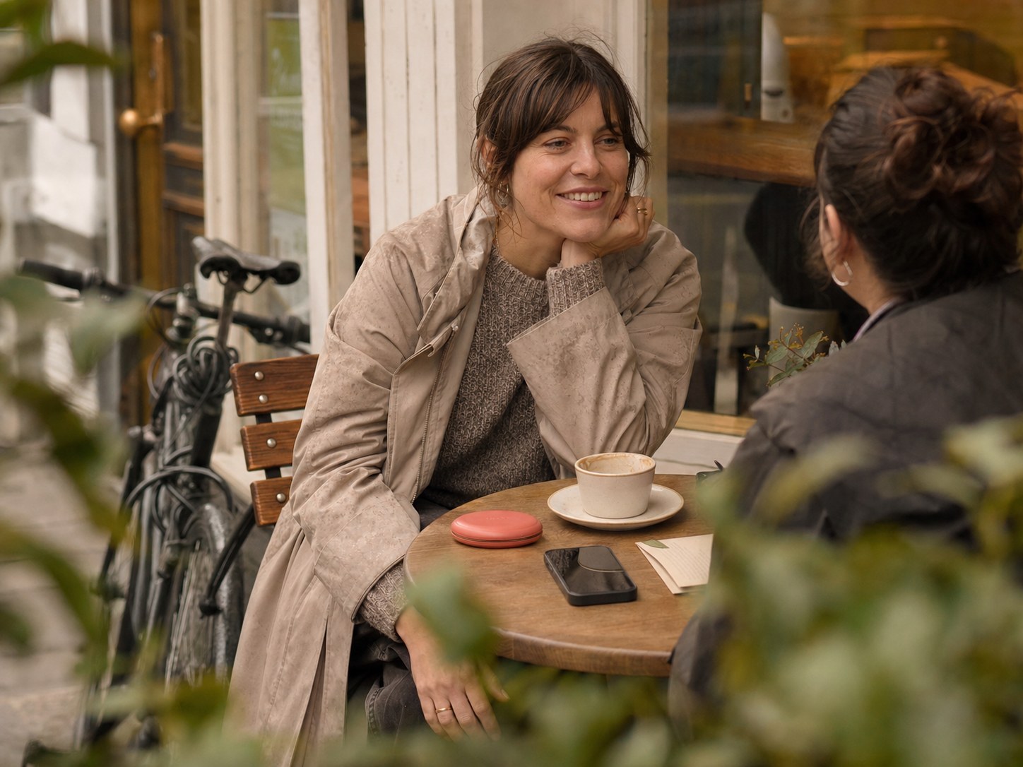 A woman with a natural smile chats outside a café beside a coral aligner case, face-down phone and folded paperwork
